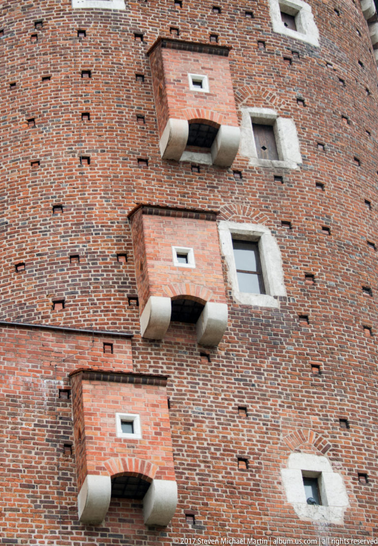 Close view of a round brick tower with small projecting openings supported by pale stone corbels.