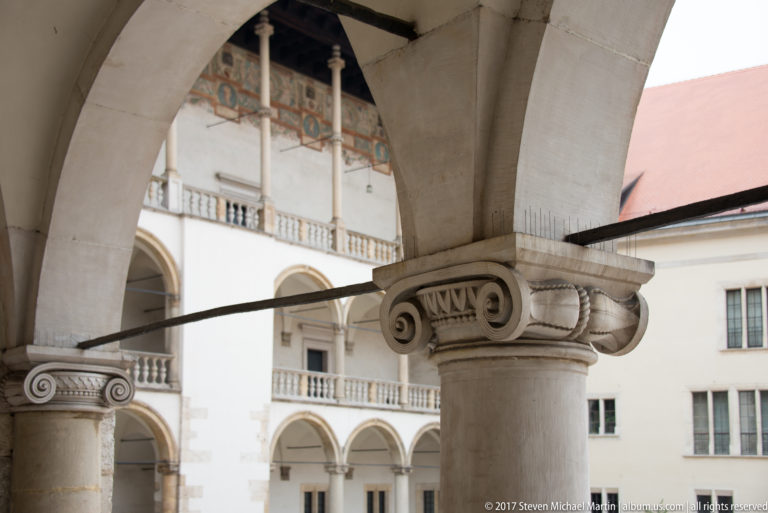 Close view of a pale stone column with volute capital beneath a Wawel courtyard arch and iron tie rod.