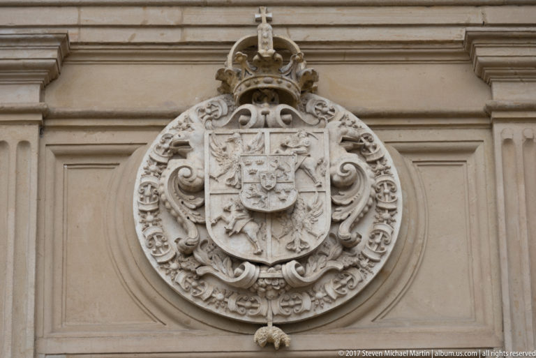 Large carved stone heraldic shield with crown, chains, and scrollwork mounted on a pale palace wall.
