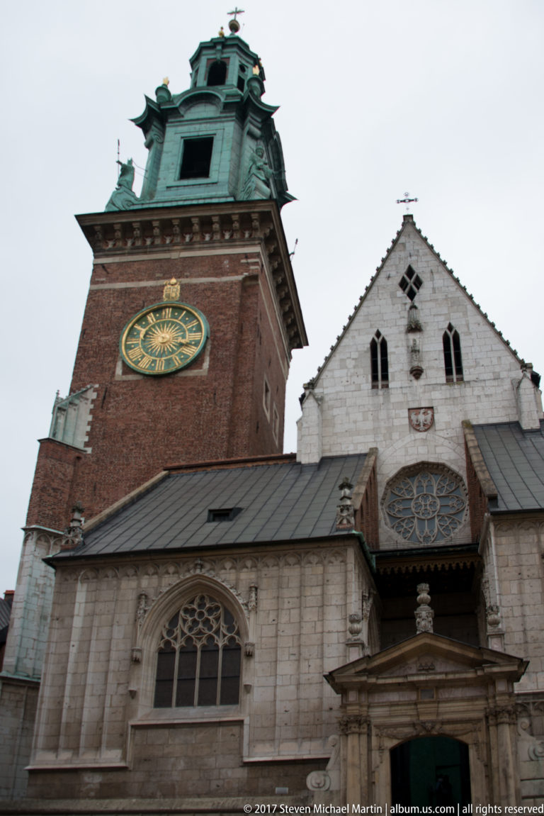 Wawel Cathedral clock tower with green copper roof beside a pale Gothic gable and traceried windows.