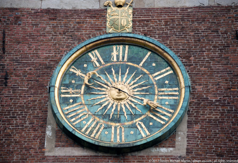 Large round clock with gilded Roman numerals and hands mounted on a red-brick tower wall.