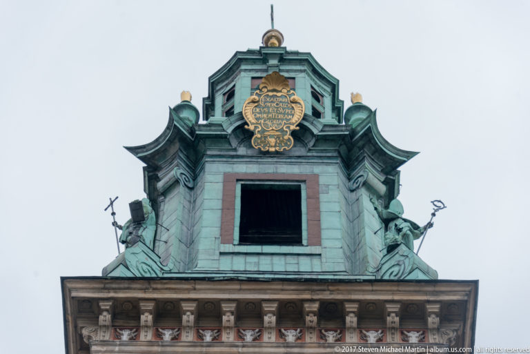 Green-patinated copper tower crown with central opening, gilded inscription plaque, and sculptural figures on both sides.