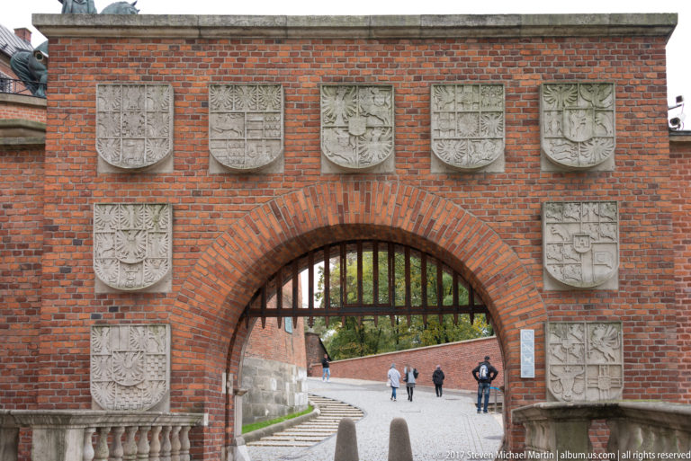 Red-brick arched gateway with iron grille surrounded by multiple carved stone coats of arms.