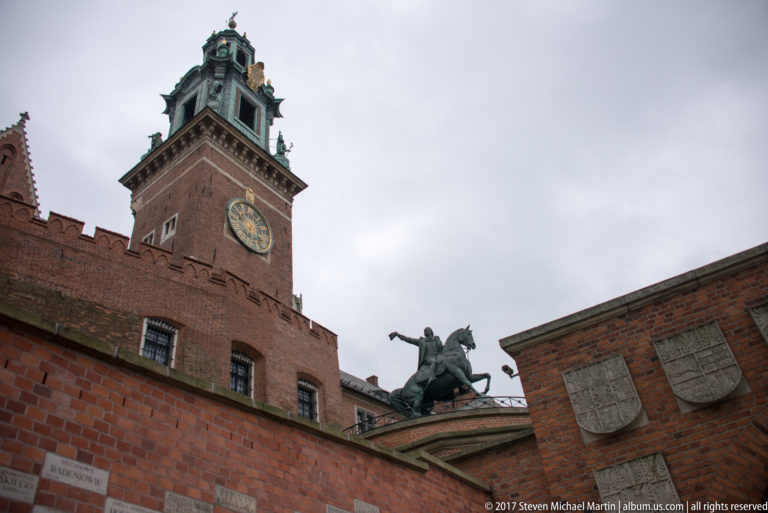 Equestrian statue above red-brick walls with Wawel's tall clock tower and heraldic plaques behind.
