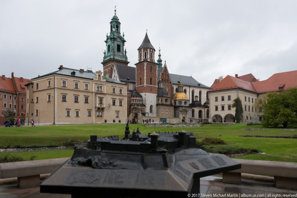 Wawel Cathedral and adjoining palace buildings seen across a lawn, with towers, spires, and a gilded dome.