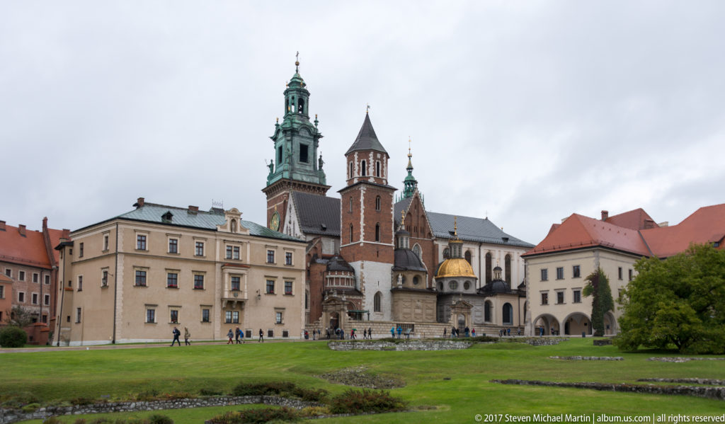 Wawel Cathedral with brick towers, green spires, and a gold dome beyond a broad green lawn.