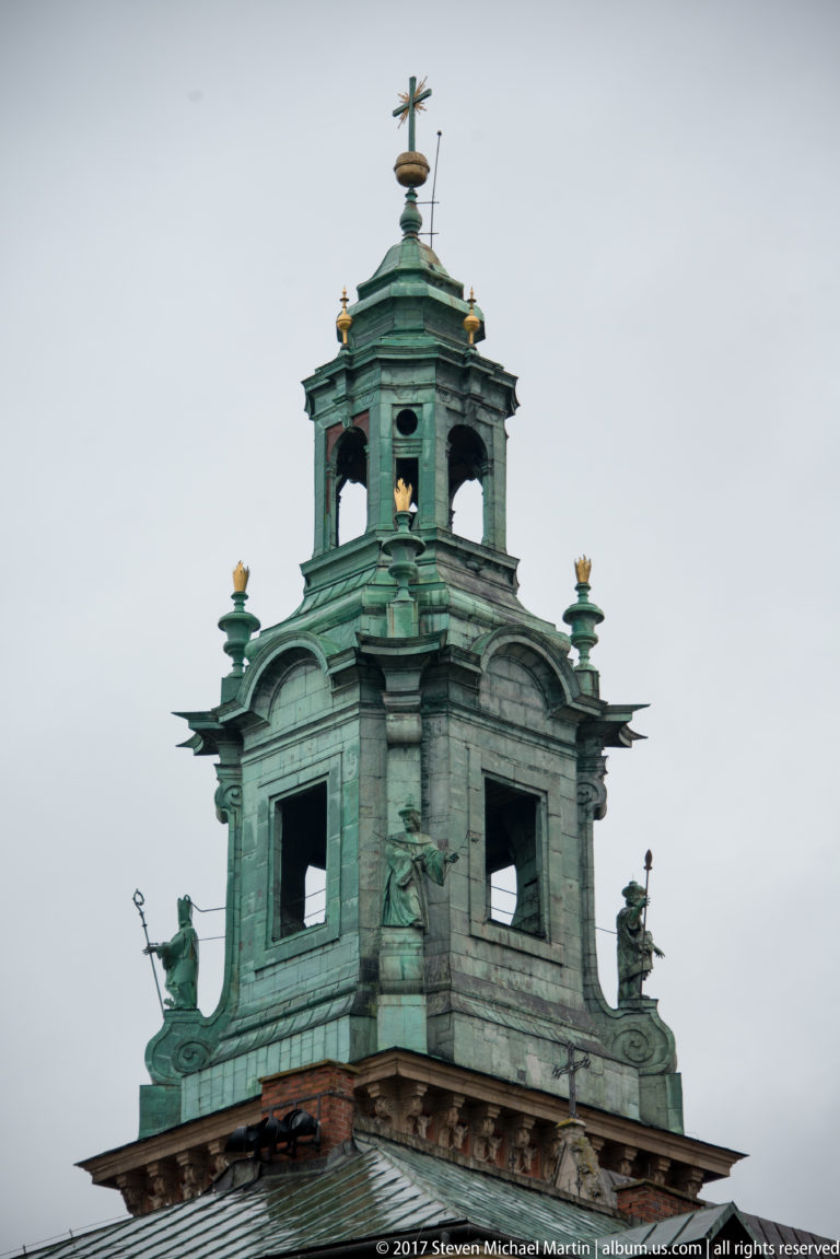 Green-patinated copper lantern with open arches, finials, and sculptural details against a gray sky.