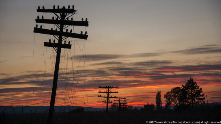 Silhouetted wooden utility poles and wires against a red-orange sunset with layered clouds.