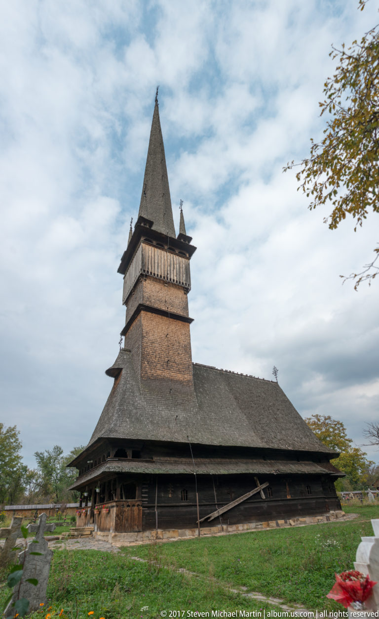 Wide side view of a dark wooden church with broad shingled roof, open belfry gallery, and steep pyramidal spire.