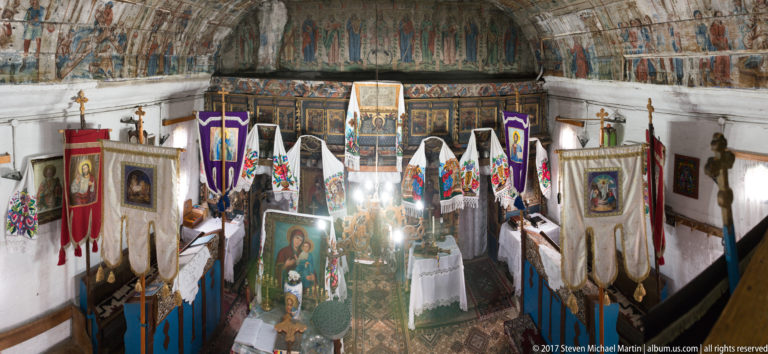 Elevated view into a painted wooden church interior filled with processional banners, icons, embroidered cloths, and devotional tables.
