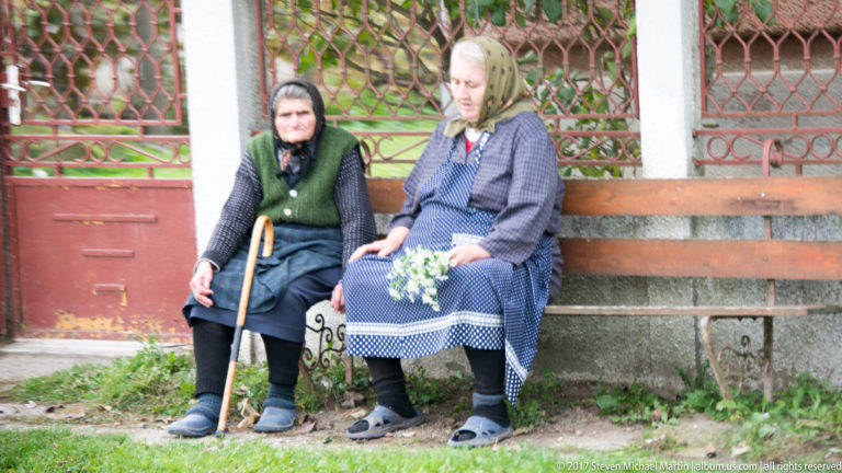 Two elderly women in headscarves seated on a bench outside a house, one with a cane and one holding flowers.