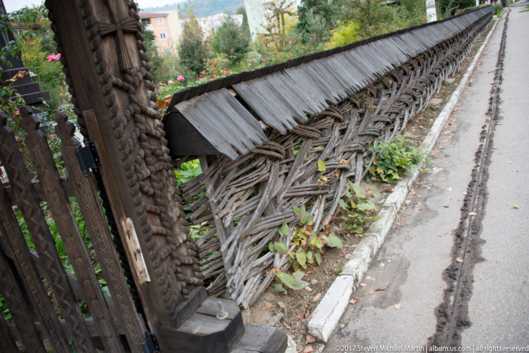 Long woven-branch roadside fence topped with a narrow pitched cap of weathered wooden shingles.