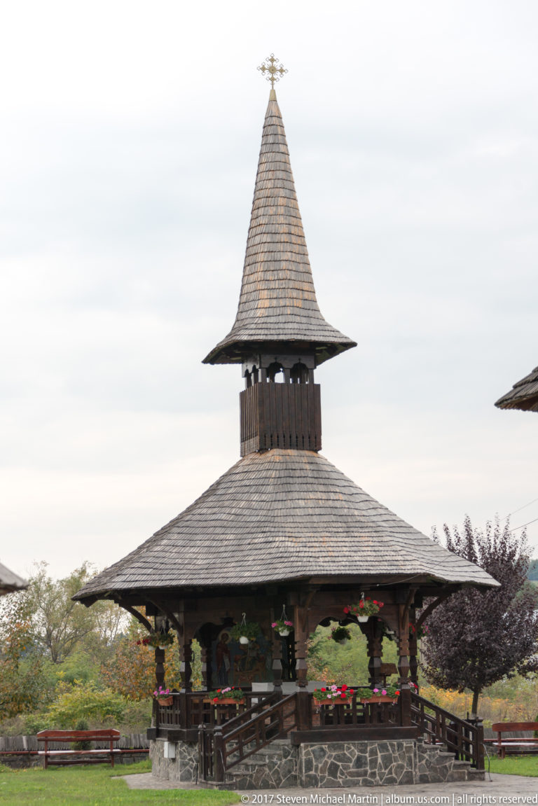 Open wooden bell pavilion with broad shingled eaves, enclosed bell stage, steep spire, cross, and flower baskets.