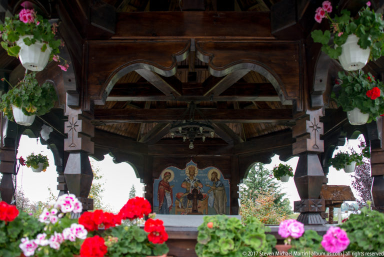 Interior of an open wooden pavilion with hanging flowers, carved posts, cross motifs, and a painted religious icon.
