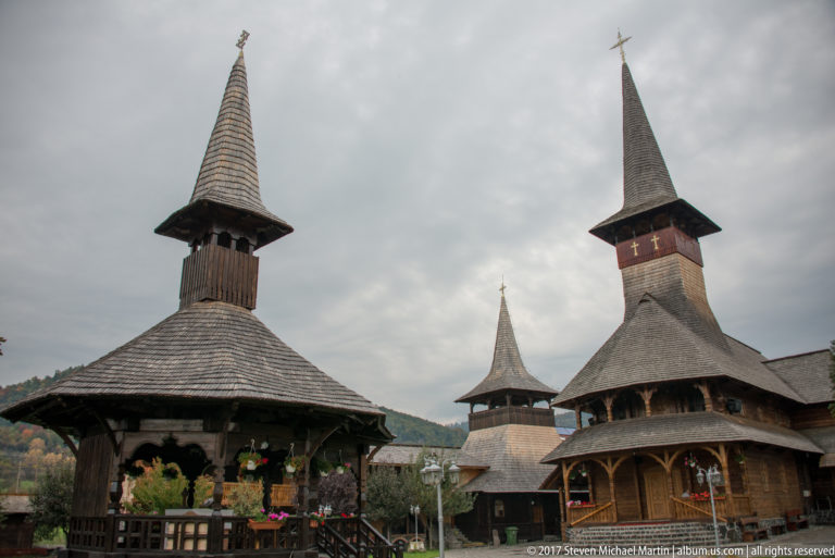 Cluster of wooden religious buildings with steep shingled roofs, tall spires, crosses, galleries, and flowers.