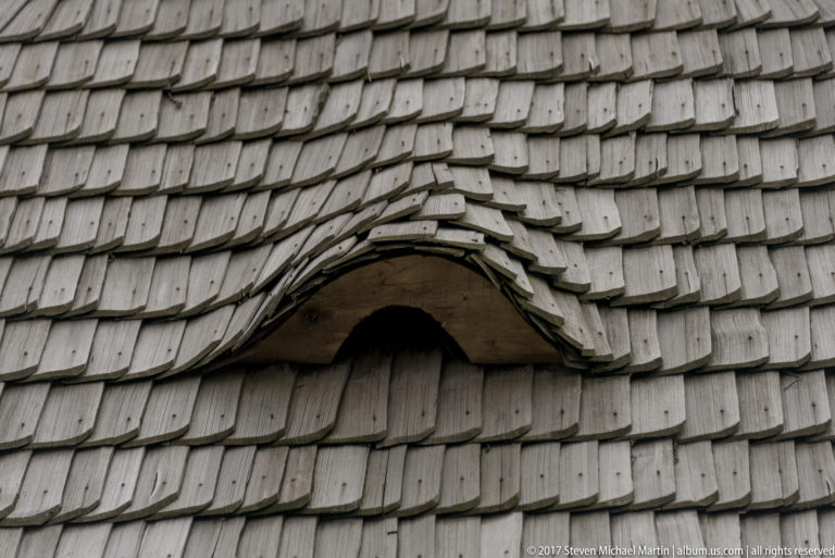 Close detail of weathered wooden shingles curving over a small eyebrow-shaped roof opening.