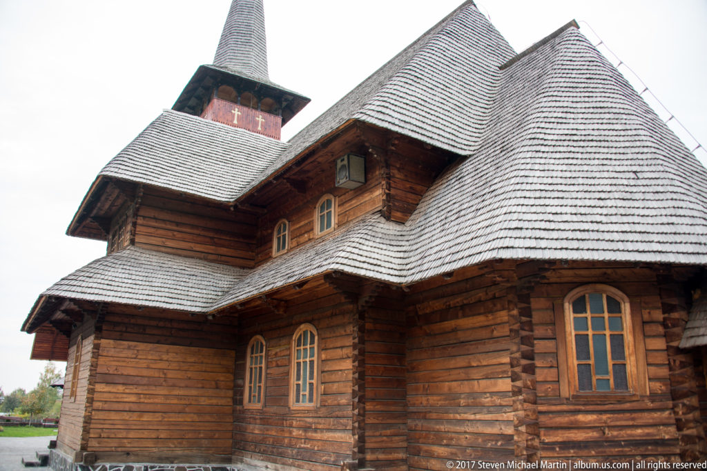 Oblique close view of a log-built wooden church with arched windows, layered shingle roofs, and a tall spire.