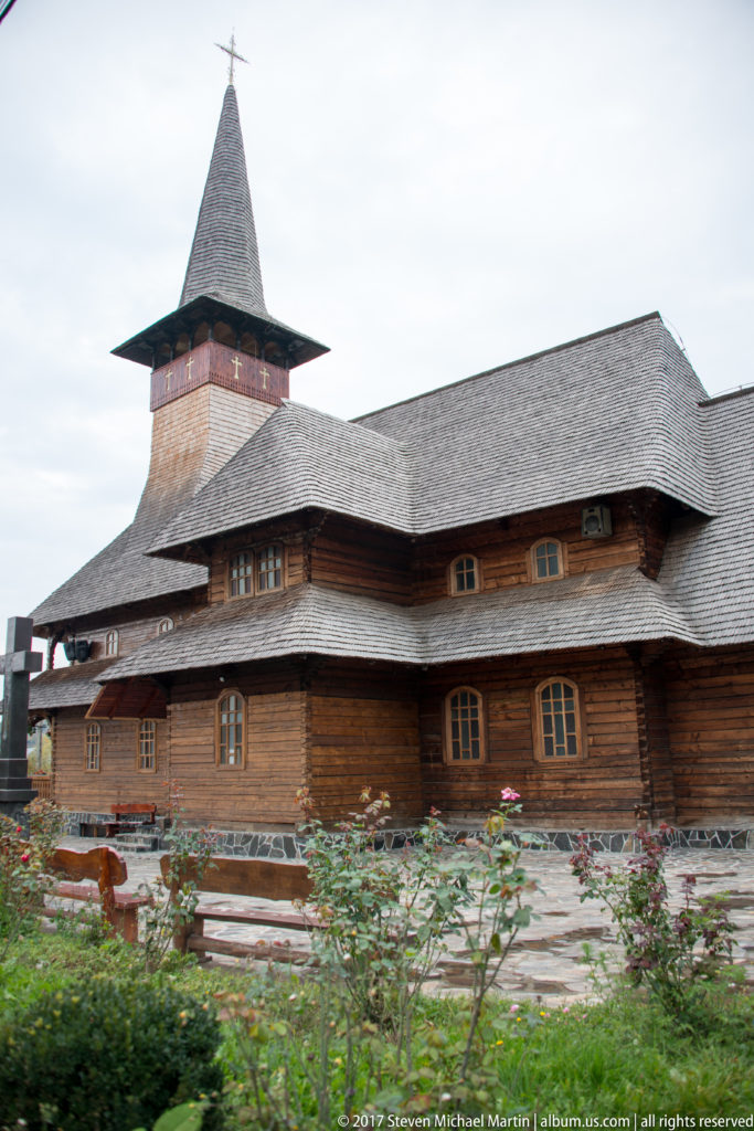 Side view of a large log-built wooden church with arched windows, broad shingled roof, tower, and flower beds.