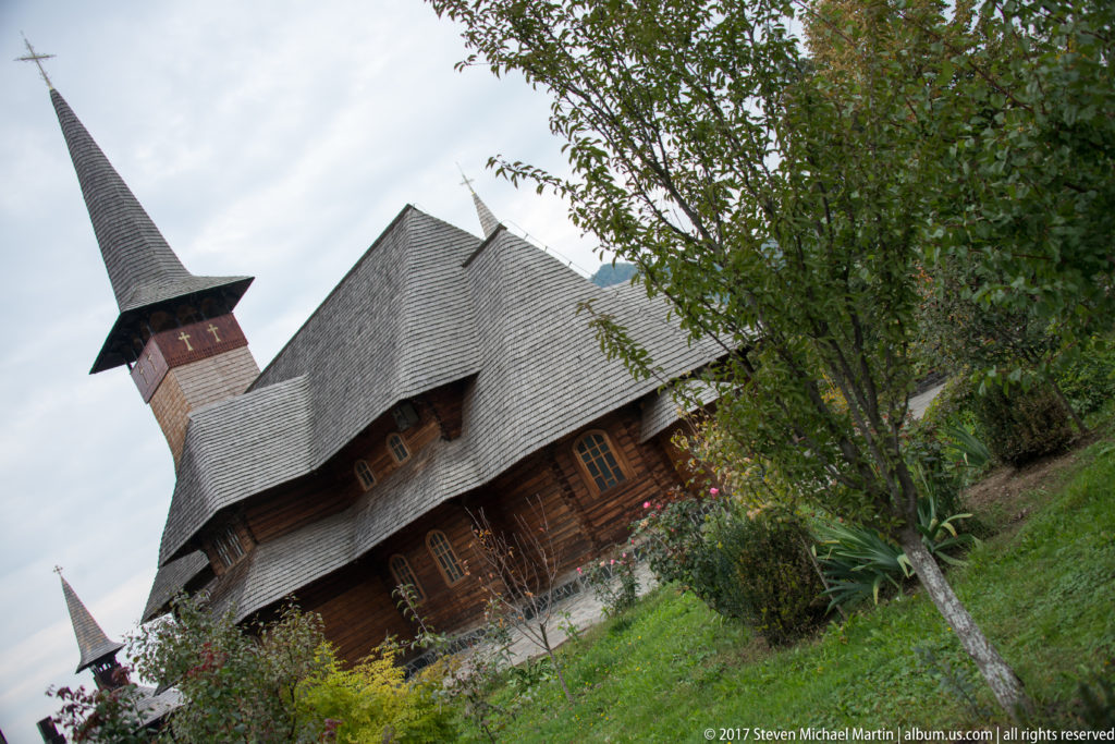 Wooden church with a tall spire and layered shingle roofs seen behind a tree on a sloping garden.