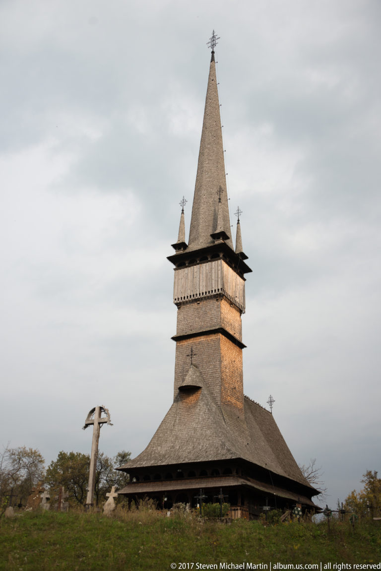 Wooden Church of Șurdești with a tall slender spire, four corner pinnacles, broad shingled roof, and nearby stone cross.