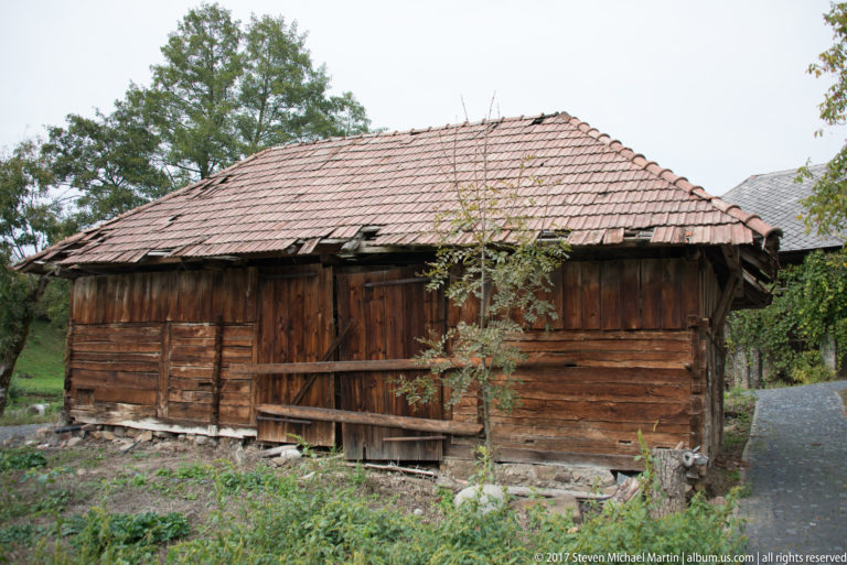 Weathered wooden rural outbuilding with log and plank walls, stone base, and a broad reddish tiled roof.