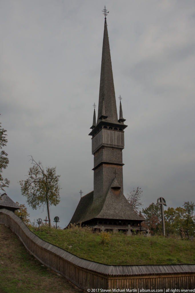 Wooden Church of Șurdești beyond a curving approach, with tall spire, corner pinnacles, shingled roof, and stone cross.