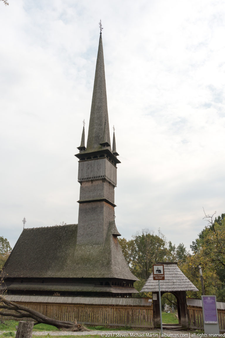 Side view of the Wooden Church of Șurdești behind a low wooden fence, with tall spire and four corner pinnacles.
