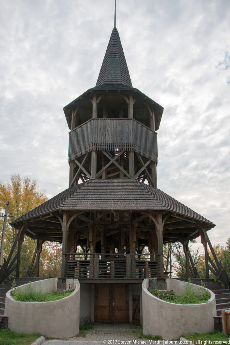 Large open timber pavilion with broad shingled roofs, exposed framing, railings, and a steep central tower.