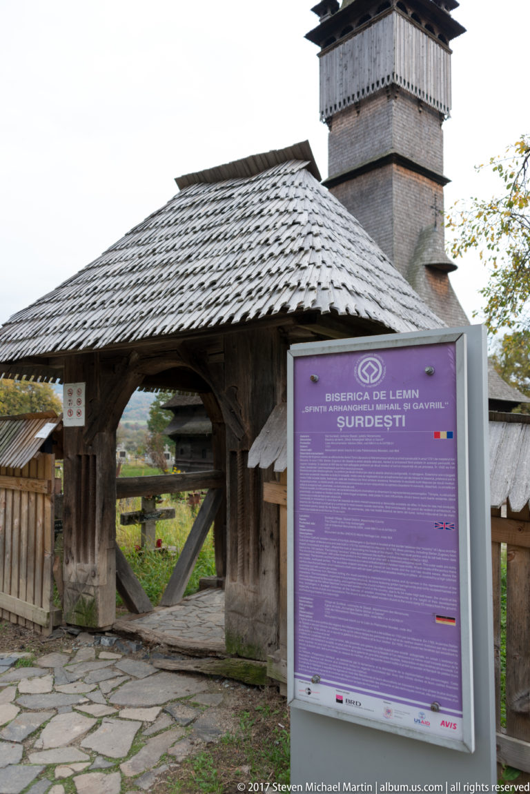 Roofed wooden churchyard gate beside a multilingual information panel identifying the wooden church at Șurdești.