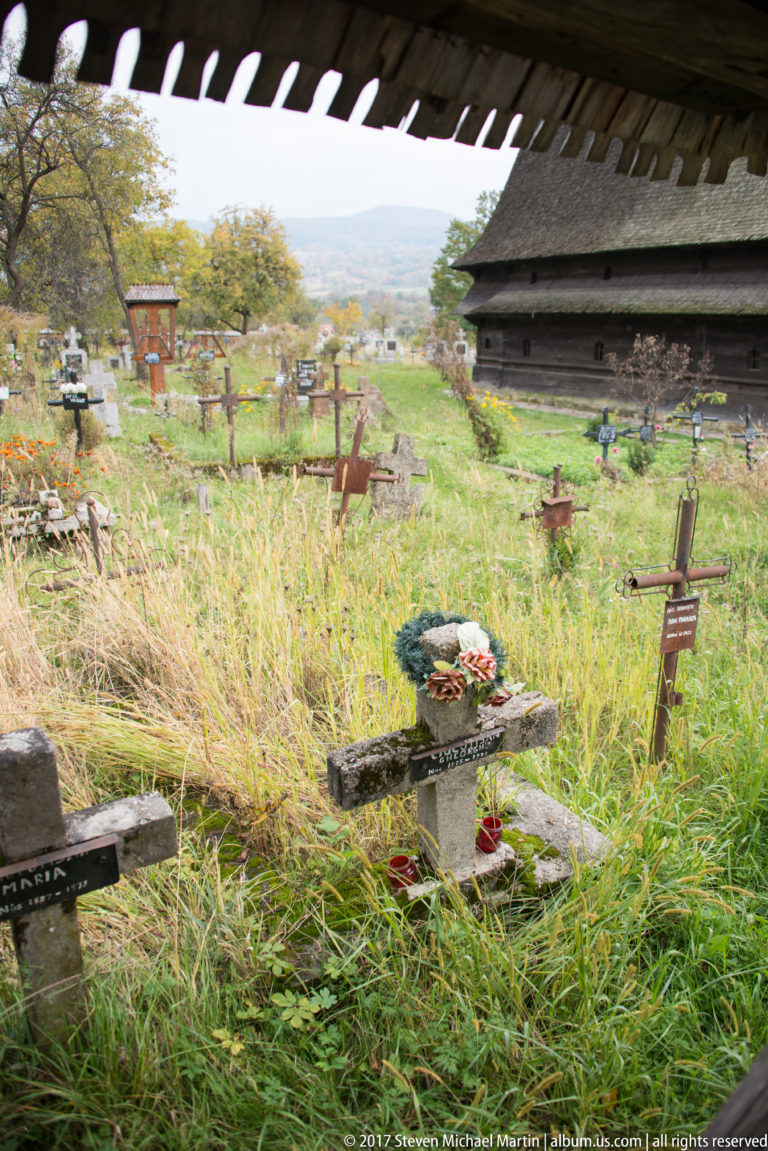 Stone and wooden grave crosses in tall grass beside the dark timber wall of the Wooden Church of Plopiș.