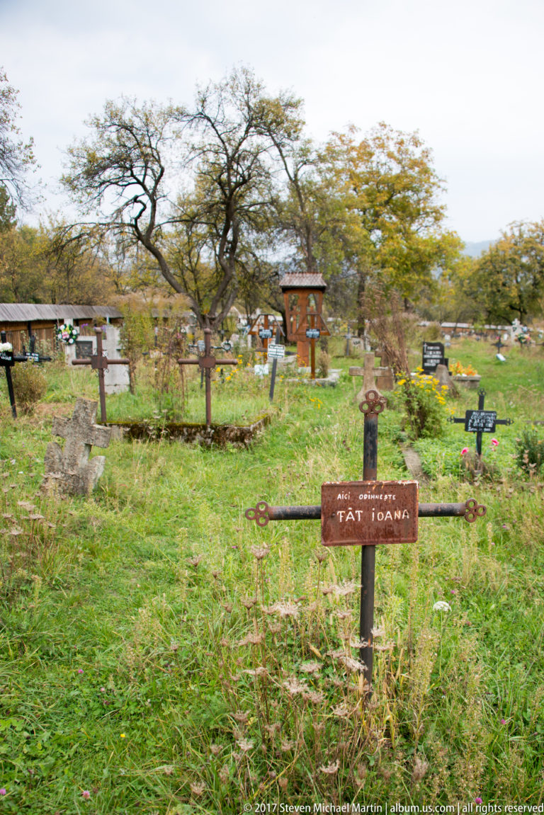 Foreground wooden grave cross with many additional crosses and trees across the Plopiș churchyard cemetery.