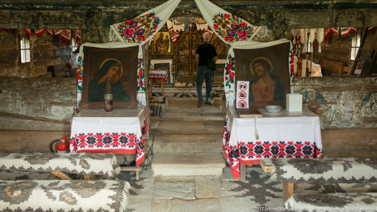 Two framed icons on white-covered tables, surrounded by embroidered textiles and rugs inside the Wooden Church of Plopiș.