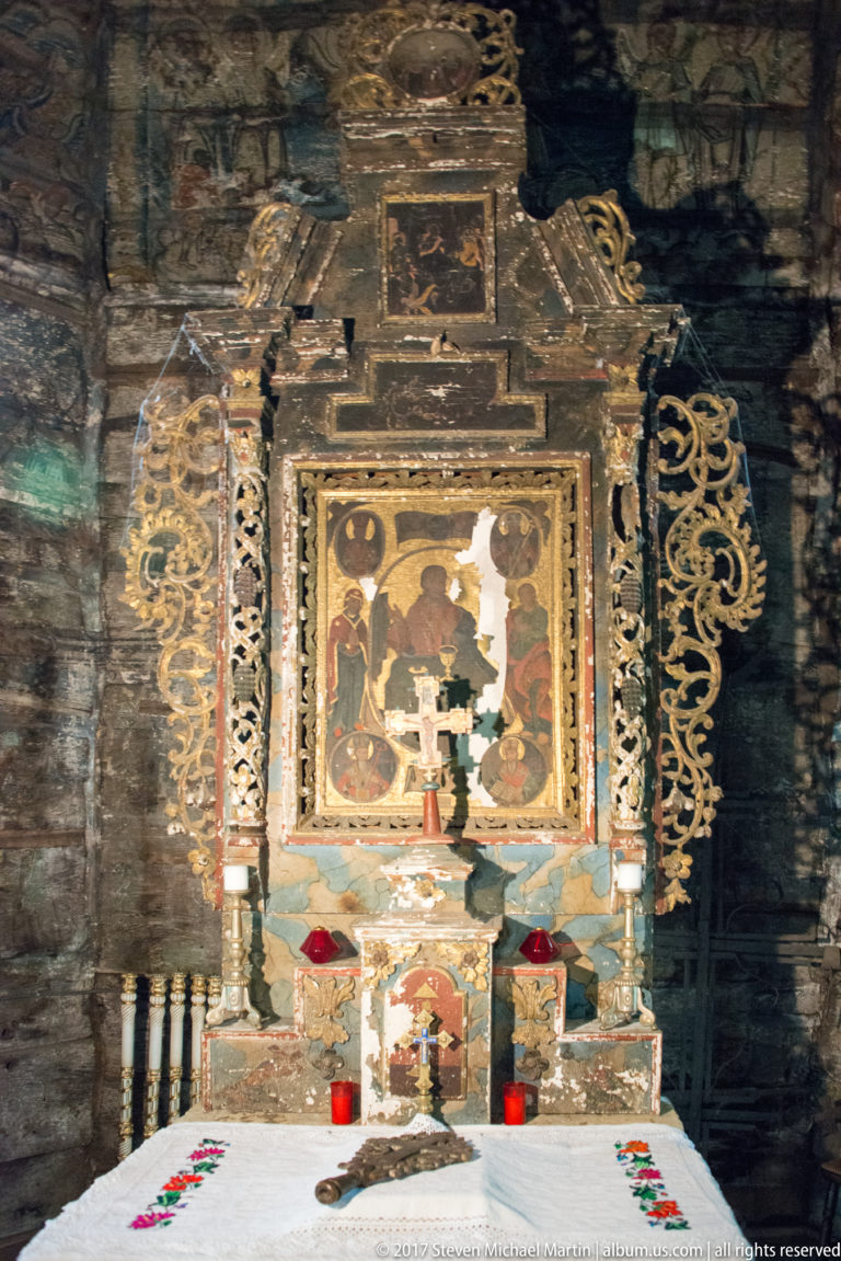 Framed devotional icon above a white-clothed side altar with candlesticks, cross, and red votive lamps.