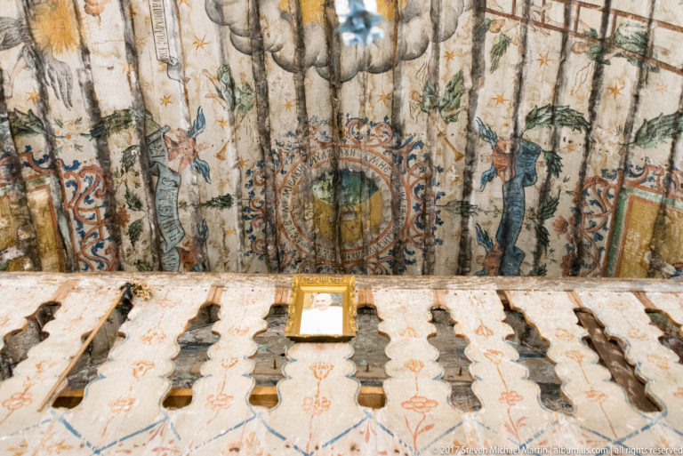Painted wooden vault above a pale carved screen with repeated baluster openings inside the Plopiș church.