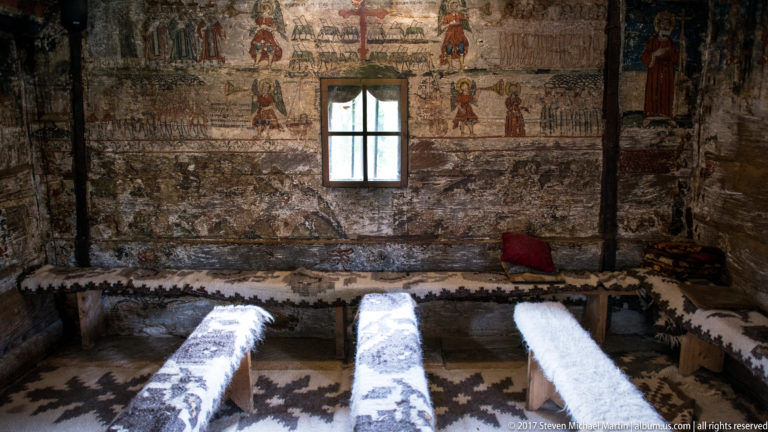 Simple benches beneath a small window and faded wall paintings inside the Wooden Church of Plopiș.