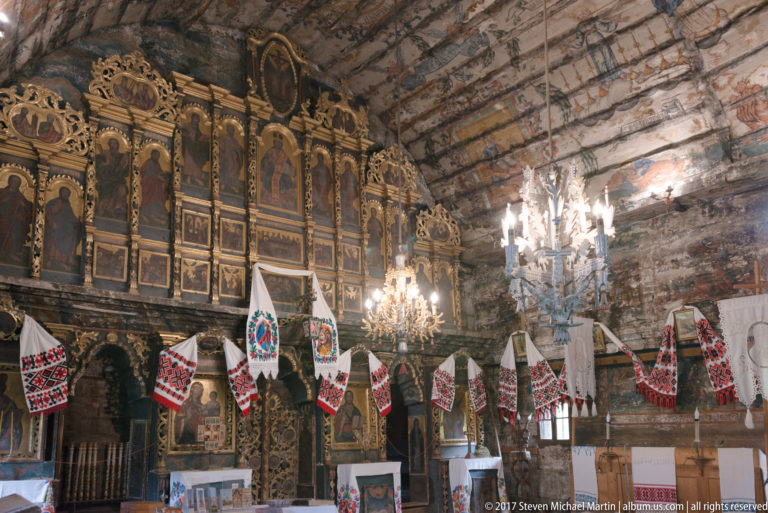 Oblique nave view toward the gilded iconostasis with chandeliers, embroidered cloths, benches, and painted timber walls.