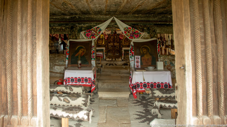 Carved wooden posts framing two devotional icons, embroidered cloths, and patterned rugs inside the Plopiș church.