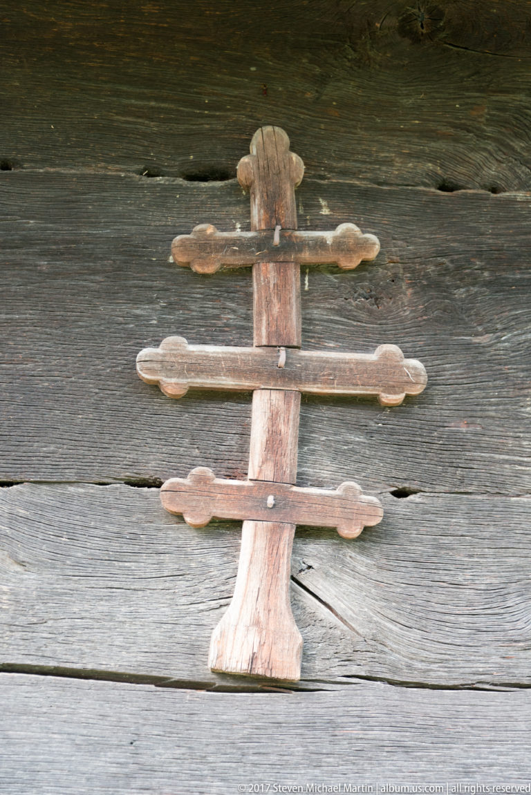 Small three-bar weathered wooden cross fixed to the dark plank wall of the Wooden Church of Plopiș.