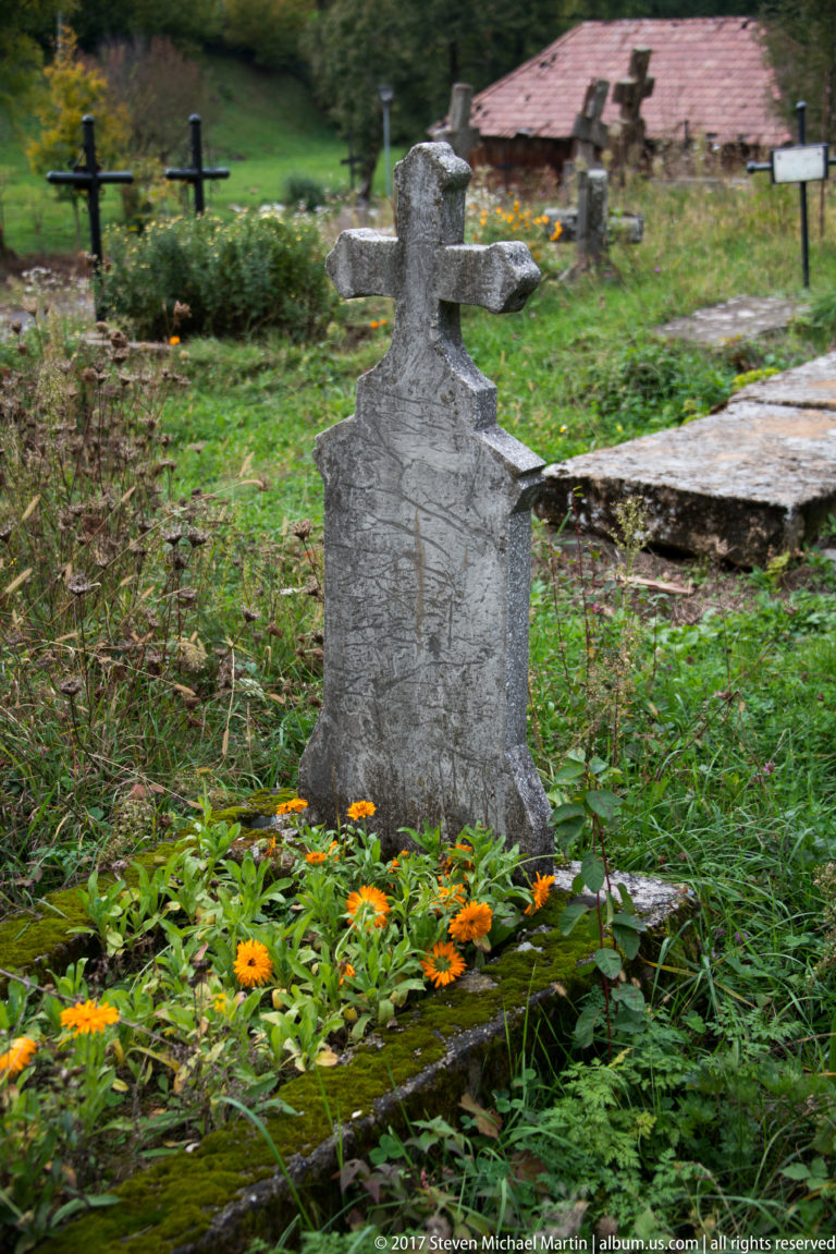 Weathered carved stone grave marker among grass and orange flowers in the Plopiș churchyard.