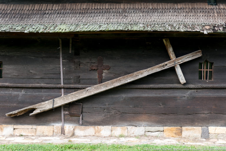 Dark timber church wall with long diagonal support beams, small cross ornaments, narrow window, and shingled eaves.