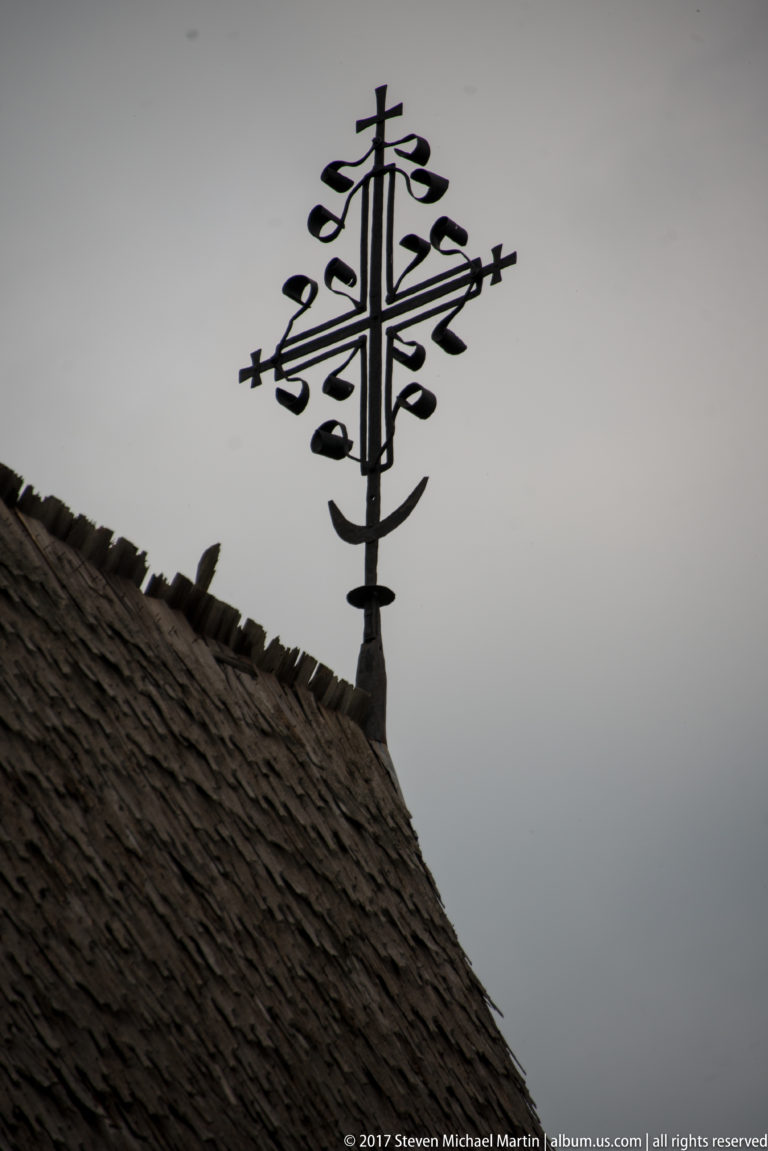 Ornate branching metal cross silhouetted above a dark wooden-shingle roof against a gray sky.