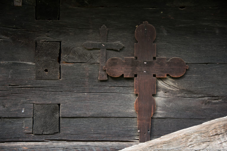 Small plain wooden cross and larger lobed three-armed cross mounted on the dark Plopiș church wall.