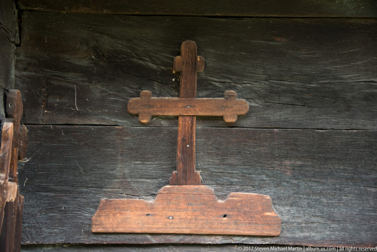 Reddish-brown lobed wooden cross with a broad base mounted against a dark timber church wall.
