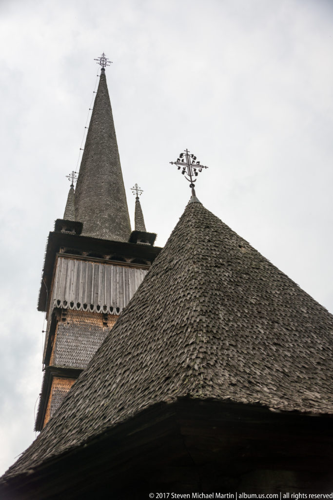 Overlapping shingled roofs, ornate roof cross, bell tower, corner pinnacles, and tall spire of the Plopiș church.