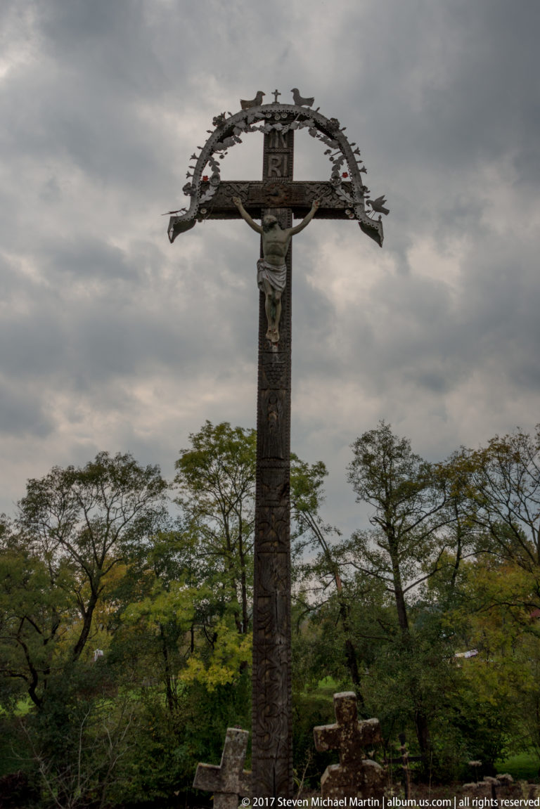 Tall outdoor wooden crucifix with figure of Christ and curved decorative canopy in the Plopiș churchyard.