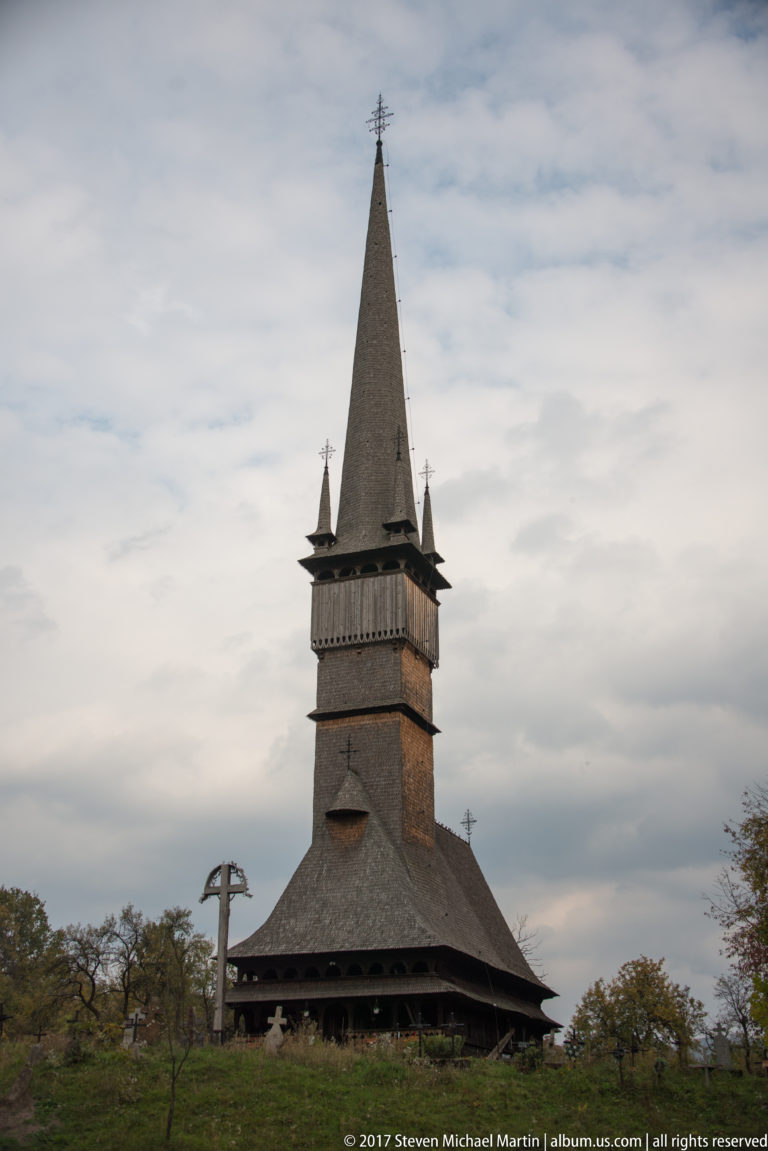 Wooden Church of Plopiș with broad shingled roof, square bell stage, corner pinnacles, tall spire, and crucifix.