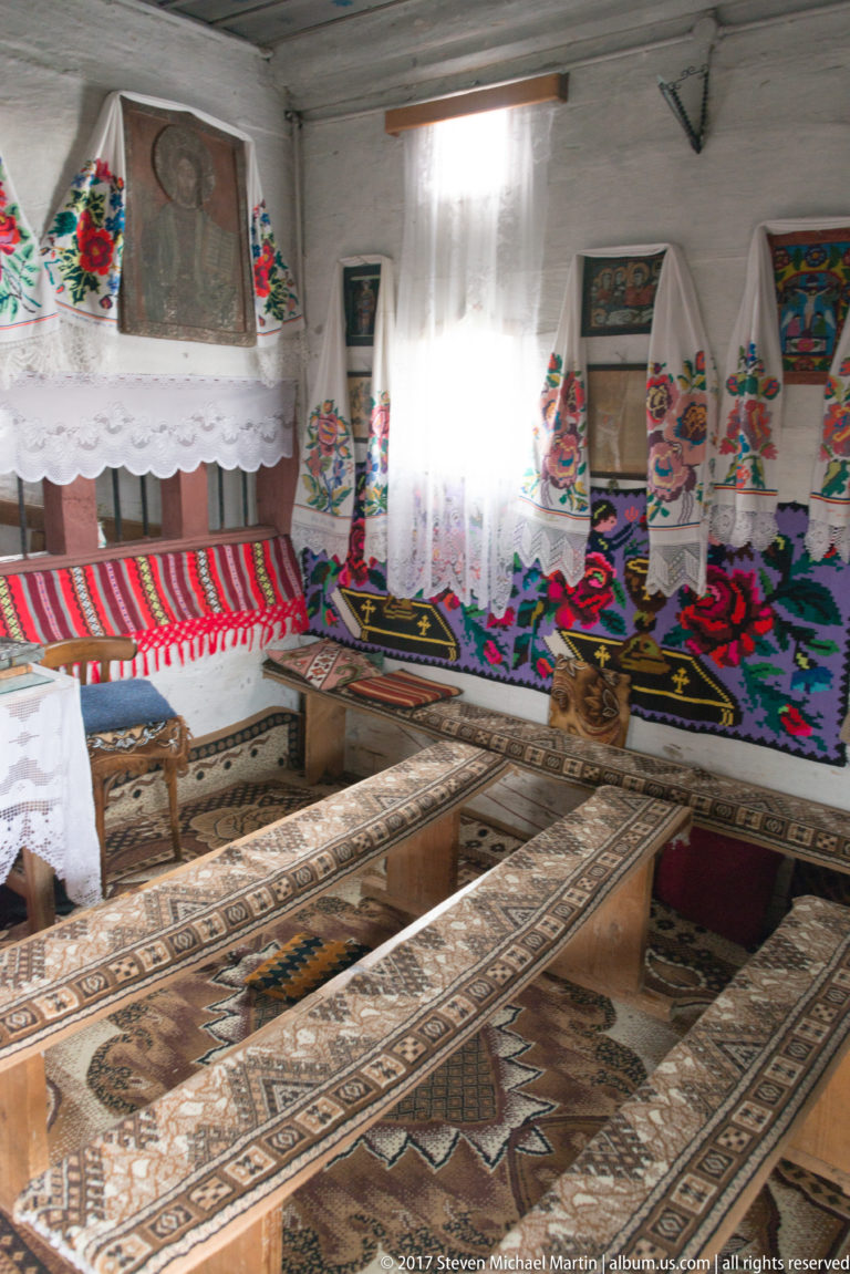 Long benches covered with patterned rugs beneath lace curtains and embroidered floral textiles inside the Plopiș church.