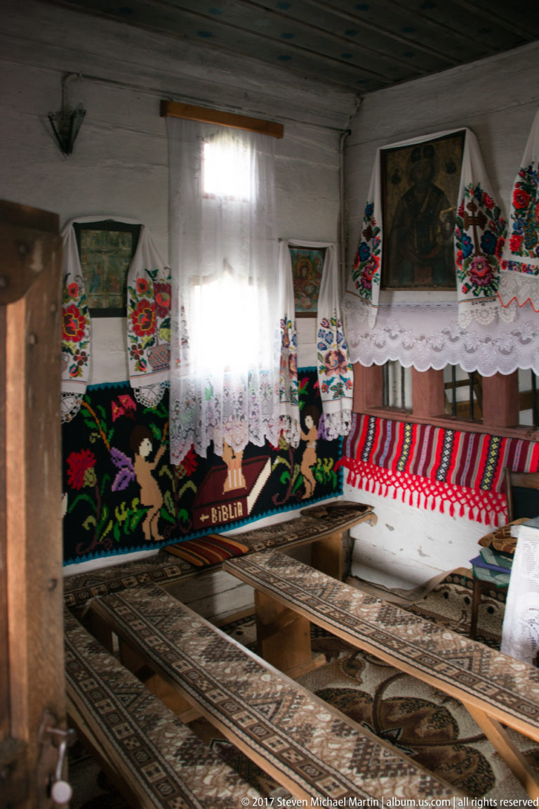 Corner interior with patterned benches, embroidered floral cloths, lace trim, framed icons, and a bright window.