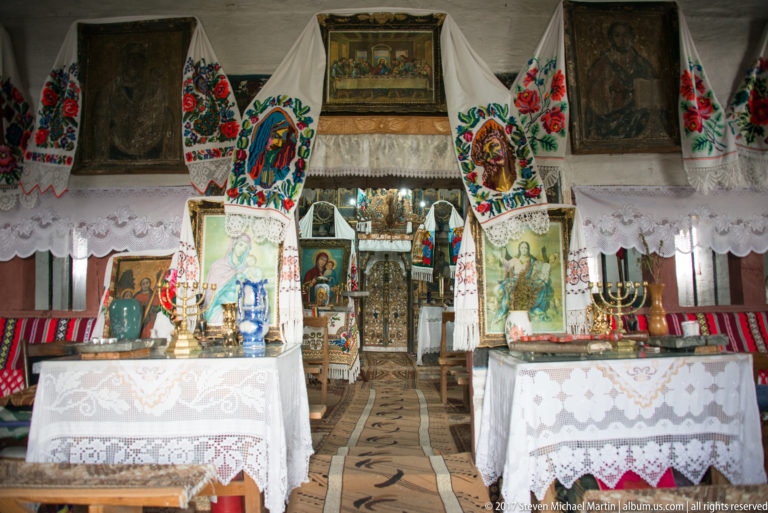 Two white-covered devotional tables with icons and candles amid embroidered textiles and religious paintings inside the Plopiș church.