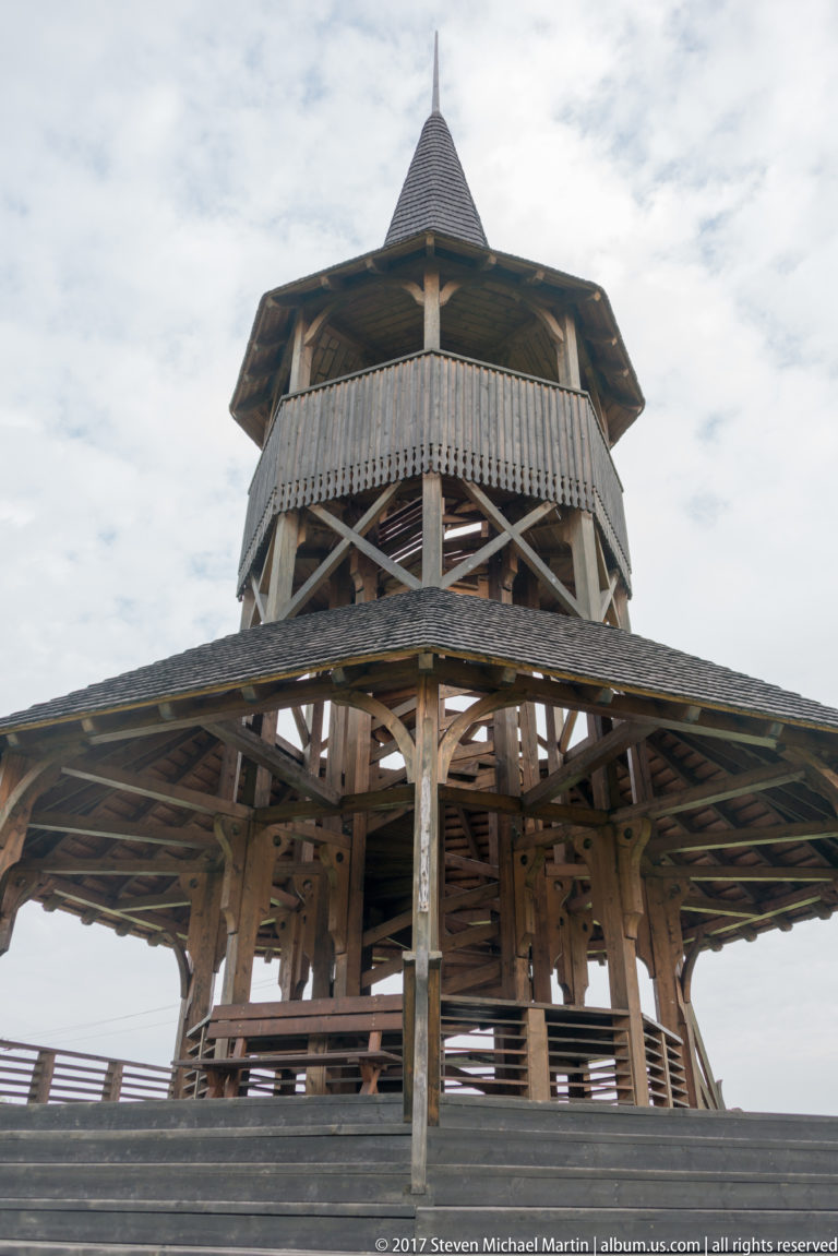 Low-angle view into exposed posts, braces, railings, and roof framing of a large wooden pavilion.